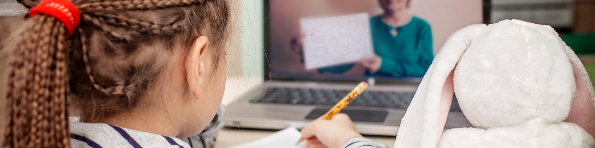 Little girl sitting with a piece of paper and a pen in front of a laptop during a video call