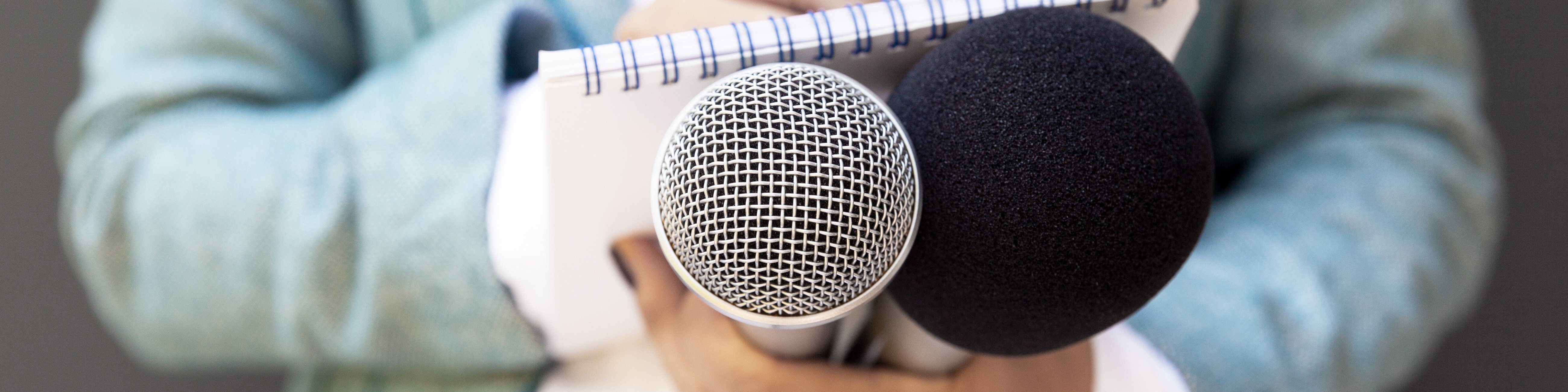 Eine Journalistin hält ein Mikrofon sowie einen Notizblock und Stift in den Händen