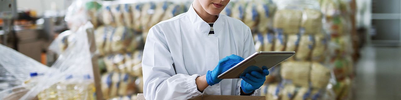 Frau im weißen Kittel, Schutzbrille und Kopfhaube steht hinter offenen Kartons mit einer Checkliste in der Hand