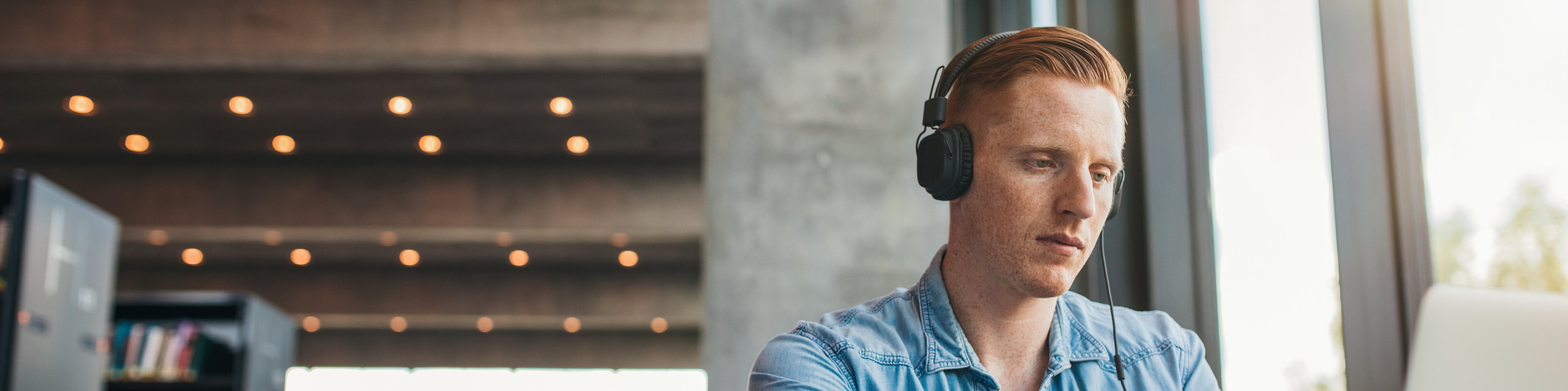 Junger Mann mit Kopfhörern sitzt an einem Laptop in einer Bibliothek